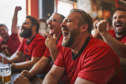 A group of men in red sports jerseys are sitting at the bar laughing and cheering for their team, watching football on TV together with friends while drinking beer photo