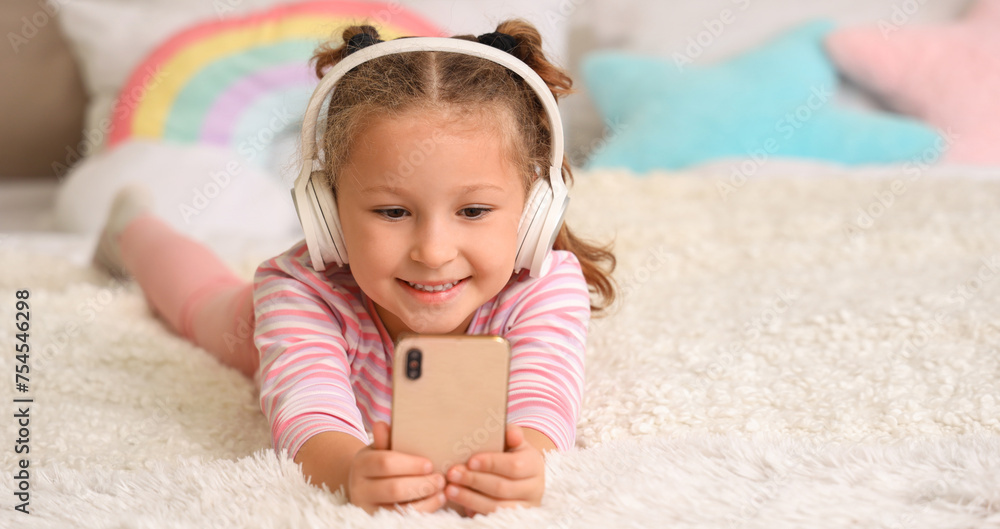 Cute little girl with headphones using mobile phone in bedroom Cute little girl with headphones using mobile phone in bedroom