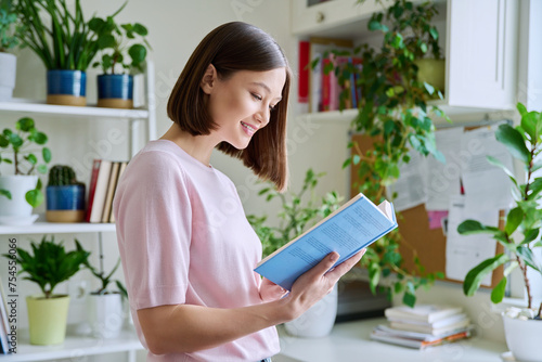 Happy smiling focused young woman reading book at home near window