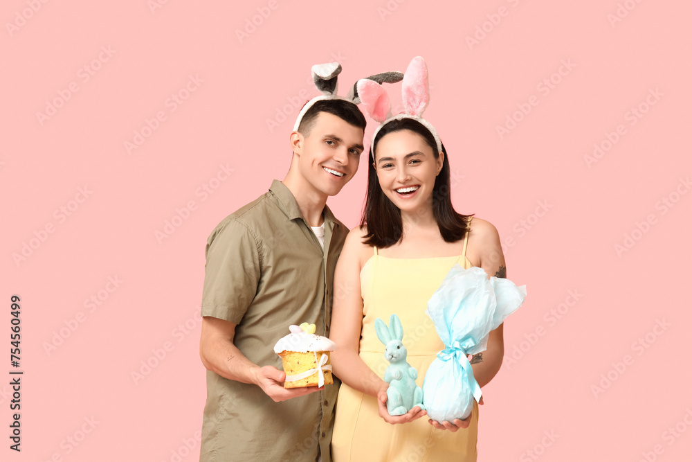 Happy young couple with Easter cake, bunny ears, gift and toy rabbit on pink background Happy young couple with Easter cake, bunny ears, gift and toy rabbit on pink background