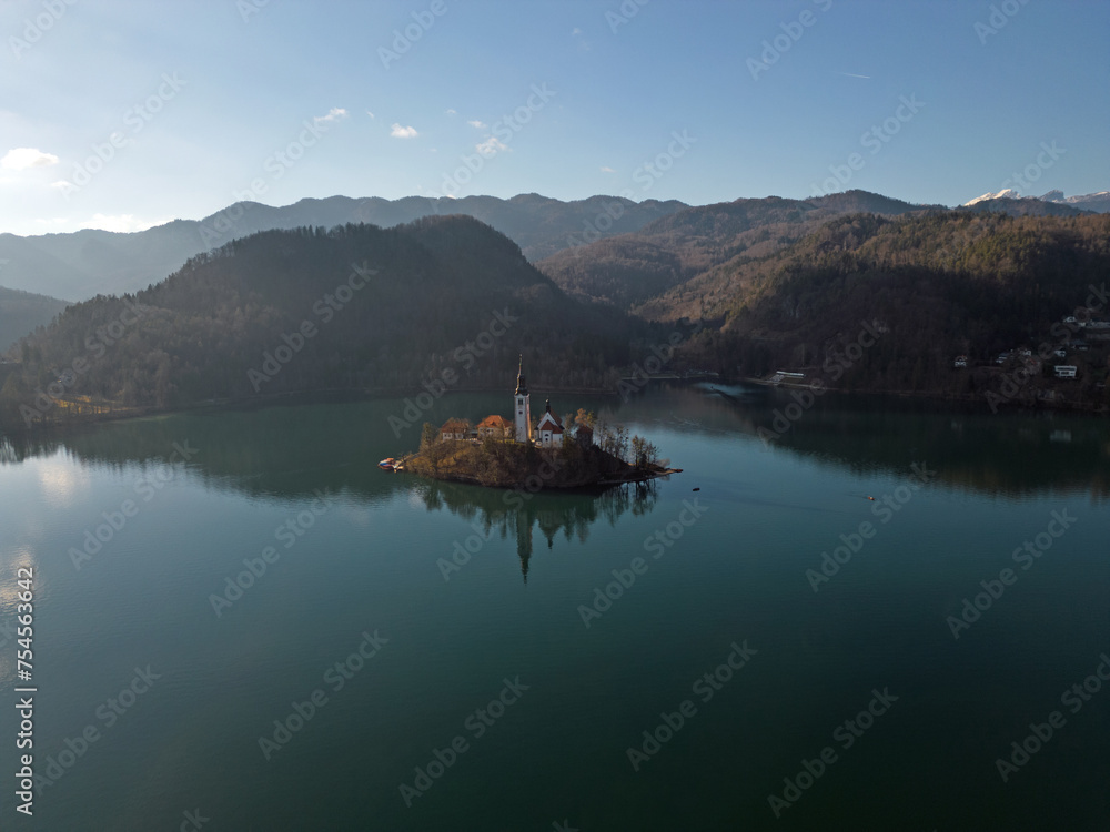 Lake Bled island with sun rays in the snowless winter aerial view. Ojstrica mountain and Julian Alps in the background. Perfect reflection of the island and the church on calm water surface.