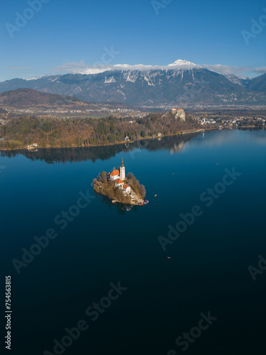 Beautiful lake Bled with island and historic church. High altitude aerial vertical view on the island and mountainous landscape on a sunny snowless winter day in Slovenia. Calm deep blue water.