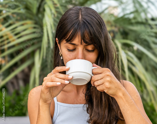 A woman is drinking coffee from a white cup with her eyes closed.