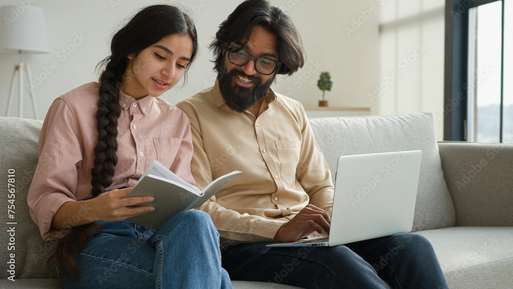 Diverse couple on couch with laptop pay bills loan payments planning household budget arabian woman wife read notes in notebook indian man husband typing data email on computer family business at home Diverse couple on couch with laptop pay bills loan payments planning household budget arabian woman wife read notes in notebook indian man husband typing data email on computer family business at home