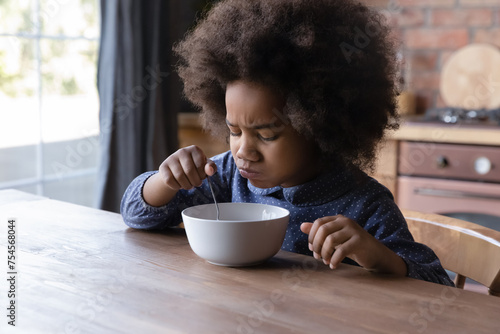 Little 6s cute African curly-haired girl sits at dining table in kitchen frowning while eats not delicious breakfast. Lack poor of appetite, healthy but not tasty monotonous food, not hungry concept photo