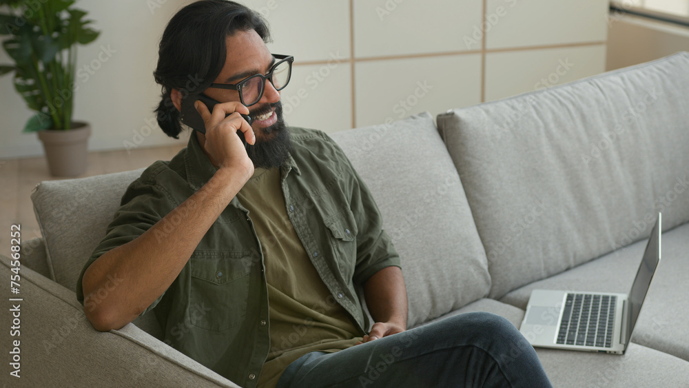 Obraz premium Muslim Arabian guy freelancer sit on couch with laptop talk mobile phone at home smiling Indian man businessman in eyeglasses speak smartphone answer call carefree converse cell business discussion