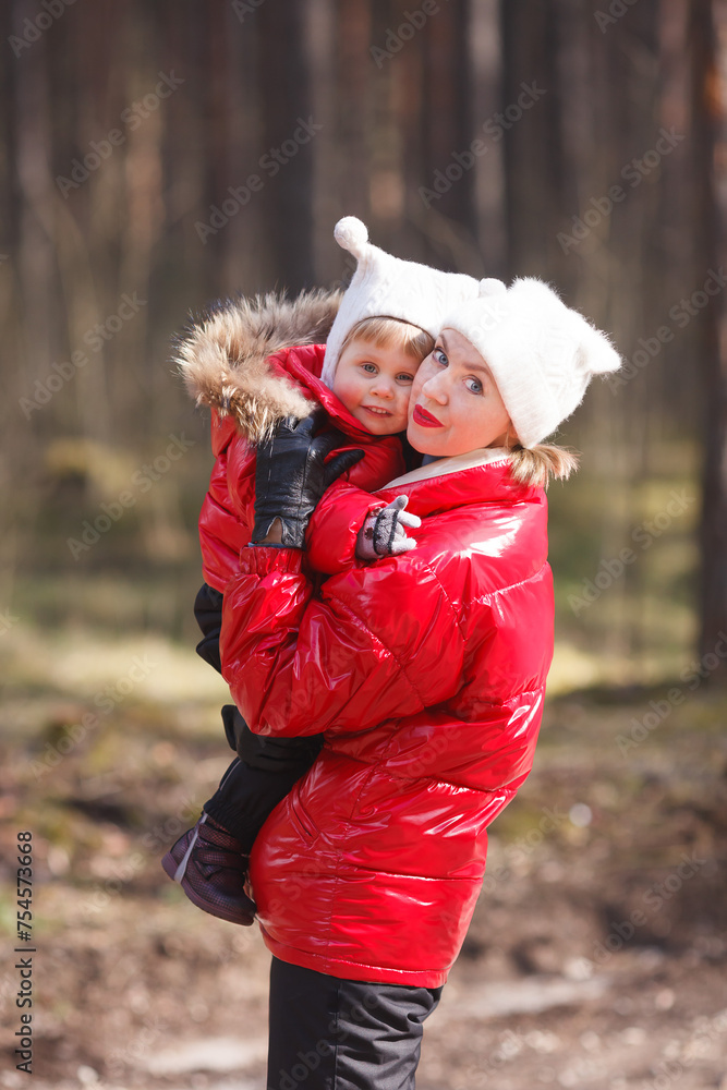 A vertical portrait of a little girl in a red winter jacket embracing her mother. Red, black and white family look winter outfit. A vertical portrait of a little girl in a red winter jacket embracing her mother. Red, black and white family look winter outfit.
