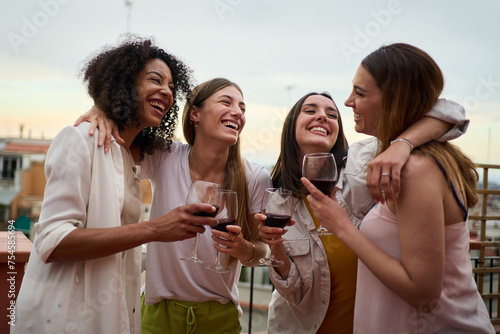 Happy group of millennial women holding glasses red wine gathered embracing celebrating summer party rooftop. Joyful young adult female friends hugging having fun together enjoying vacation outdoors #754585694