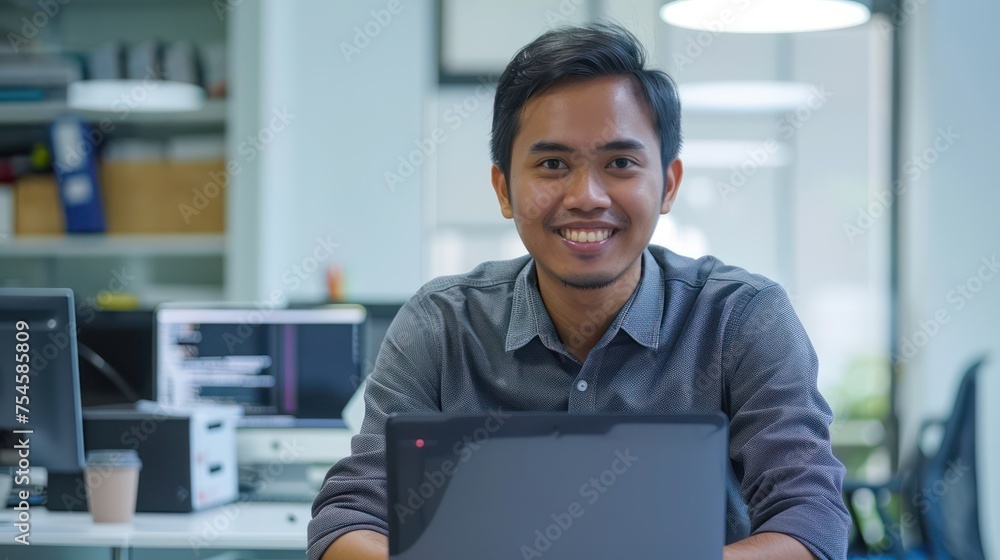 Fototapeta premium An Asian male employee smiling as he works on a laptop in the office, making eye contact with the camera.