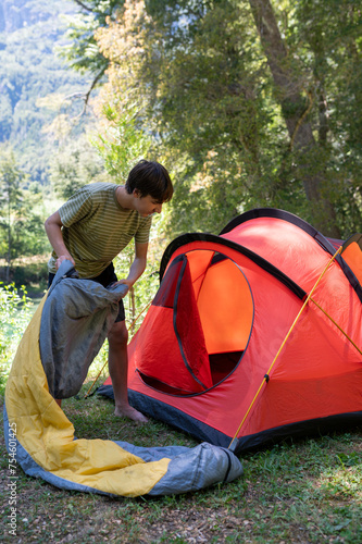 A young boy arranges his sleeping bag inside the tent, which is installed in the middle of nature between rivers and mountains.