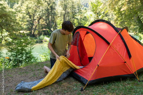 A young man arranges his camping tent and sleeping bag at the campsite during his summer walk through the mountains of southern Argentina. Surrounded by green, rivers and mountains. photo