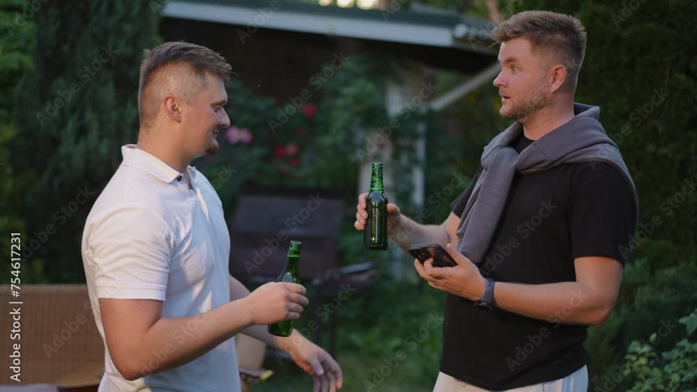 Slow motion. A man looks at the screen of a mobile phone, standing in the fresh air. A friend approaches a man with two glass bottles of beer in his hands and treats him. The man is immensely happy Slow motion. A man looks at the screen of a mobile phone, standing in the fresh air. A friend approaches a man with two glass bottles of beer in his hands and treats him. The man is immensely happy