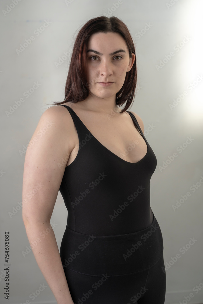 Fototapeta premium Young brunette womain in black tank top poses befoe a white wall in soft light
