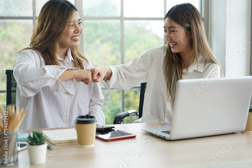 Two young Asian women roommates working at home with laptops and hand bump together with friends smiling, happy at successful online business apartment.