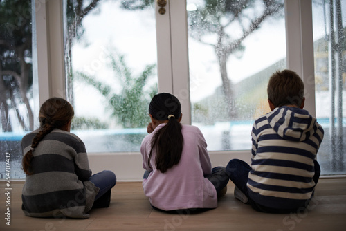 Rain, window and sad child on a floor unhappy, upset or annoyed by bad weather at home. Glass, disappointed and sibling kids in a living room frustrated by storm, cold or unexpected winter disaster