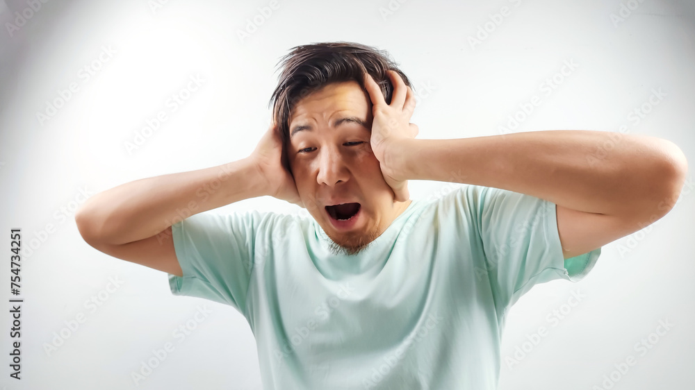 Portrait of handsome Asian man in blue t-shirt, holding head with both hands angry and screaming being stressed on isolated white background Portrait of handsome Asian man in blue t-shirt, holding head with both hands angry and screaming being stressed on isolated white background