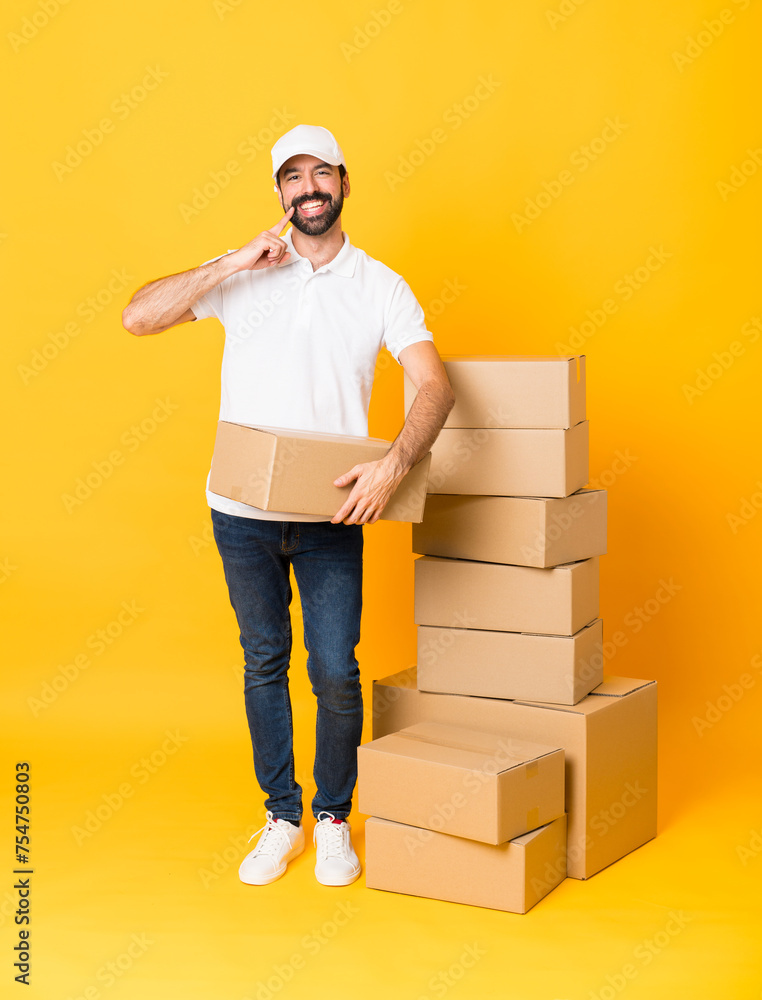 Full-length shot of delivery man among boxes over isolated yellow background smiling with a happy and pleasant expression Full-length shot of delivery man among boxes over isolated yellow background smiling with a happy and pleasant expression