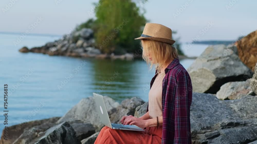 Blonde woman wearing hat sitting on the rocky beach while working on a laptop. Freelance, remote work on vacation. Blonde woman wearing hat sitting on the rocky beach while working on a laptop. Freelance, remote work on vacation.