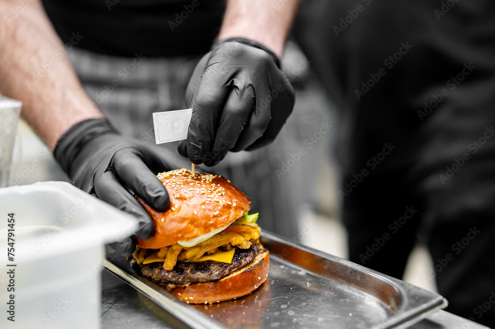 A chef in black gloves assembles a juicy burger with fresh ingredients on a metal tray in a professional kitchen setting