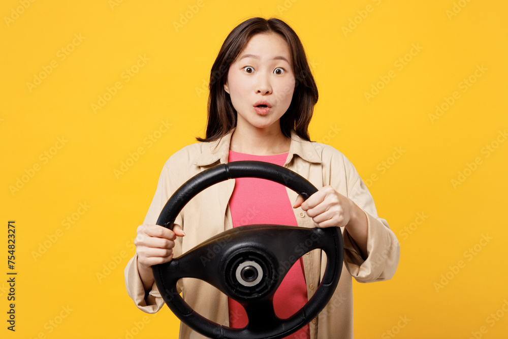 Young amazed woman of Asian ethnicity she wear pink t-shirt beige shirt pastel casual clothes hold steering wheel driving car isolated on plain yellow background studio portrait. Lifestyle portrait. Young amazed woman of Asian ethnicity she wear pink t-shirt beige shirt pastel casual clothes hold steering wheel driving car isolated on plain yellow background studio portrait. Lifestyle portrait.