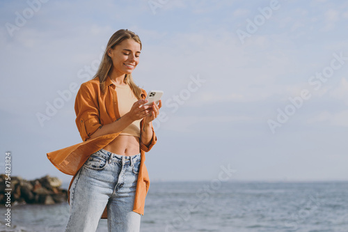 Side bottom view young smiling calm woman she wear orange shirt casual clothes hold in hand use mobile cell phone walk on sea ocean sand shore beach outdoor seaside in summer day. Lifestyle concept