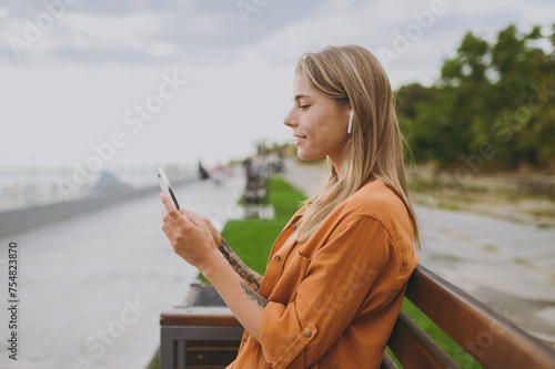 Side view young woman wear orange shirt casual clothes use mobile cell phone listen music in earphones sit on bench walk rest relax in spring green city park outdoor on nature Urban lifestyle concept