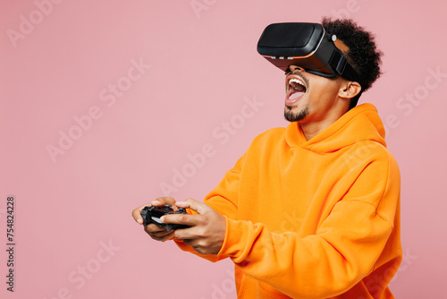 Side view young man of African American ethnicity in yellow hoody casual clothes hold in hand play pc game with joystick console watch in vr headset pc gadget isolated on plain light pink background