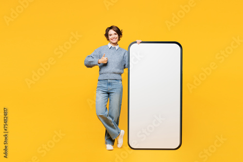 Full body young woman wears grey knitted sweater shirt casual clothes big huge blank screen mobile cell phone smartphone with area show thumb up isolated on plain yellow background. Lifestyle concept.