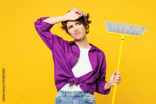 Young sad tired exhausted woman wears purple shirt casual clothes do housework tidy up hold broom put hand on head sweating isolated on plain yellow background studio portrait. Housekeeping concept.