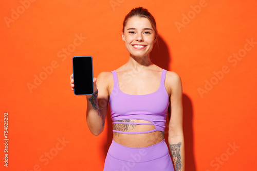 Young fitness trainer sporty woman sportsman wear purple top clothes spend time in home gym hold use blank screen mobile cell phone isolated on plain orange background. Workout sport fit abs concept.