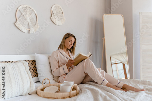 pretty smiling woman relaxing at home on bed in morning in pajamas reading book