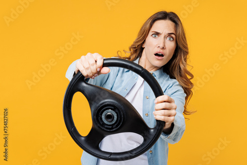 Young shocked sad scared woman she wearing blue shirt white t-shirt casual clothes hold steering wheel driving car look camera isolated on plain yellow background studio portrait. Lifestyle concept.