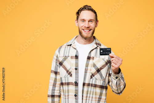 Young smiling happy Caucasian man he wear brown shirt casual clothes hold in hand mock up of credit bank card look camera isolated on plain yellow orange background studio portrait. Lifestyle concept.