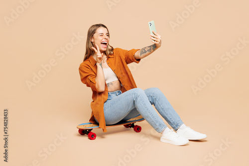 Full body young woman wear orange shirt casual clothes sits on pennyboard skateboard do selfie shot on mobile cell phone show v-sign isolated on plain pastel light beige background. Lifestyle concept.