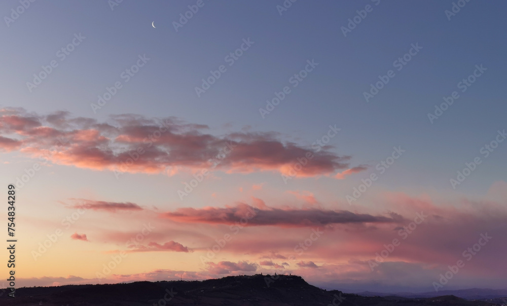La Luna all’alba resta ancora nel cielo sopra le nuvole rosa sulla colline mentre il sole sorge dal mare