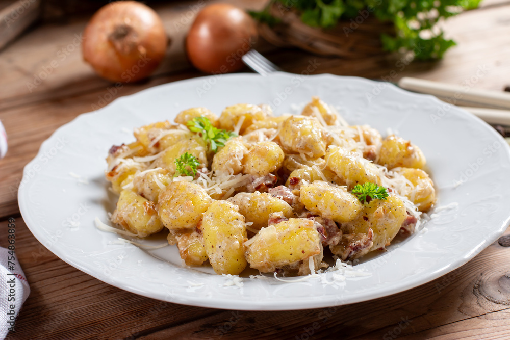 Gnocchi (small potato balls) with bacon in creamy sauce in a white plate . Close up view. Lunch table background, fork, garlic. Traditional Italian cuisine. Gnocchi (small potato balls) with bacon in creamy sauce in a white plate . Close up view. Lunch table background, fork, garlic. Traditional Italian cuisine.