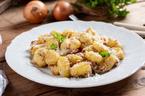 Gnocchi (small potato balls) with bacon in creamy sauce in a white plate . Close up view. Lunch table background, fork, garlic. Traditional Italian cuisine.