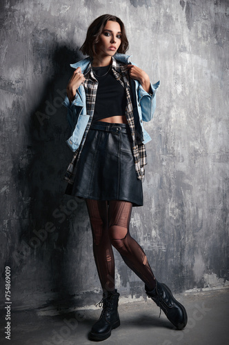 Indoor portrait of lovely grunge (rock) girl standing at wall. Cool informal model, dressed in a jean jacket, checkered shirt, leather skirt and torn pantyhose
