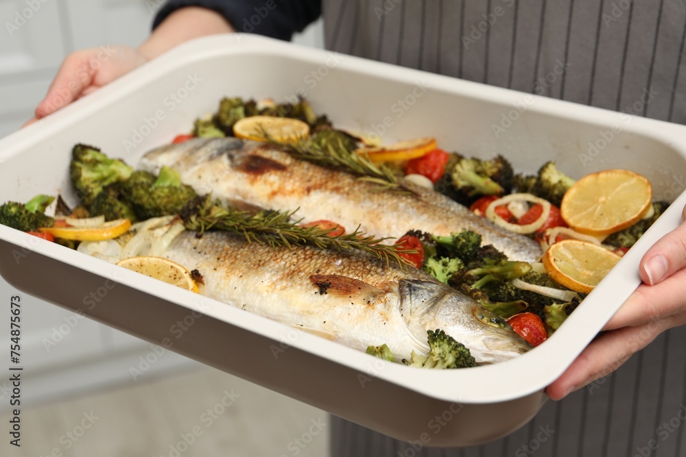 Woman holding baking dish with delicious fish and vegetables indoors, selective focus Woman holding baking dish with delicious fish and vegetables indoors, selective focus