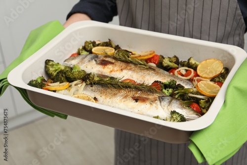 Woman holding baking dish with delicious fish and vegetables indoors, closeup