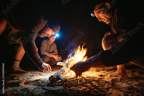 Three men gathered around a small campfire on a beach at night, two with headlamps