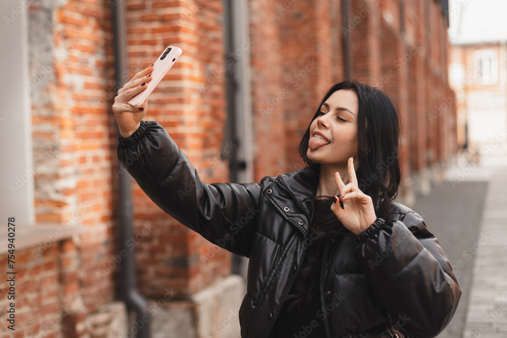 Happy young woman photographing herself using her mobile phone. Caucasian female taking selfie with her smart phone at city. Woman show peace sign and tongue out, talk by video call, wear black puffer