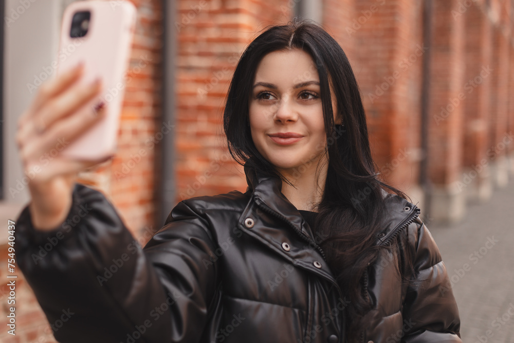 Pensive brunette woman make selfie posing on street brick building background. Outdoor shot of happy hippie lady make video call, smiling. Girl wear black puffer jacket raises her hand hold camera. Pensive brunette woman make selfie posing on street brick building background. Outdoor shot of happy hippie lady make video call, smiling. Girl wear black puffer jacket raises her hand hold camera.