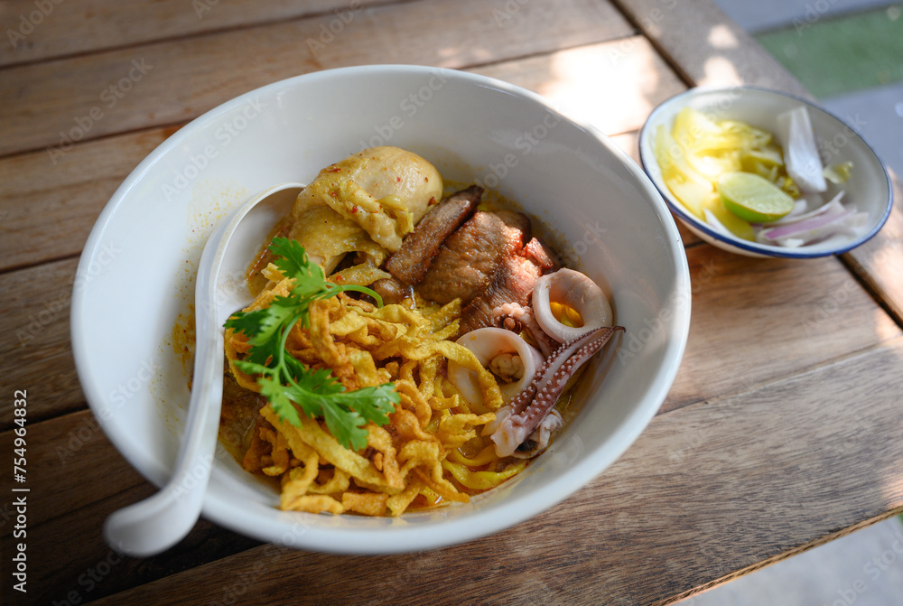 A bowl of Khao Soi, a Northern Thai coconut curry noodle soup with chicken, seafood, and crispy noodles, served with lime and pickled vegetables.
