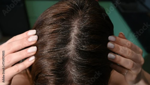 a woman touches her hair, looking at early gray hair. Parting of dark brown female hair with gray roots. close-up