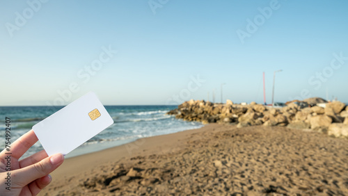 In a woman's hand a white bank card against a background of beach and sea. © Jūlija