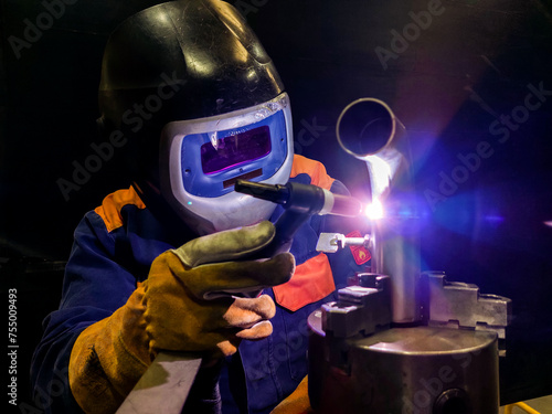 A professional welder performing TIG (Tungsten Inert Gas) welding on stainless steel. The welder is equipped with protective gear, including a welding helmet, heat-resistant gloves