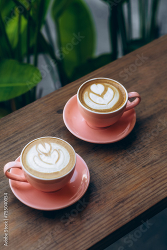 vertical shot of two coral-colored coffee mugs with intricate latte art, resting on a rustic wooden table against a soft green backdrop