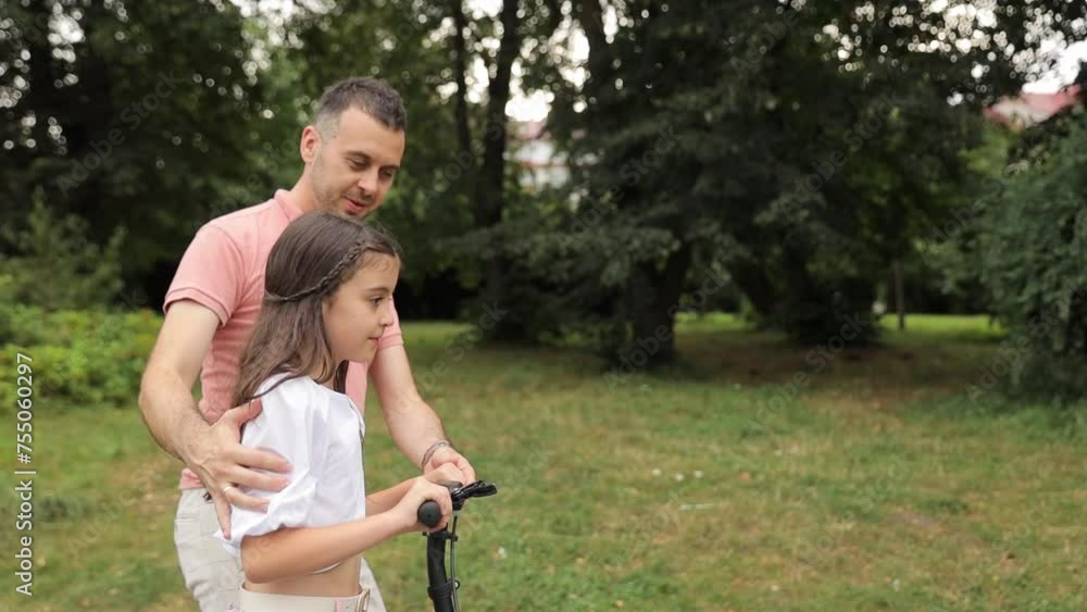 A man is pushing a little girl on a scooter through a park, surrounded by lush green grass, trees, and a peaceful natural landscape. A man is pushing a little girl on a scooter through a park, surrounded by lush green grass, trees, and a peaceful natural landscape.