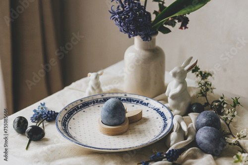 Happy Easter! Stylish easter egg in bunny figurine on vintage plate and spring flowers on linen, rustic table setting. Natural painted blue eggs and hyacinth blooms. Modern minimal still life © sonyachny